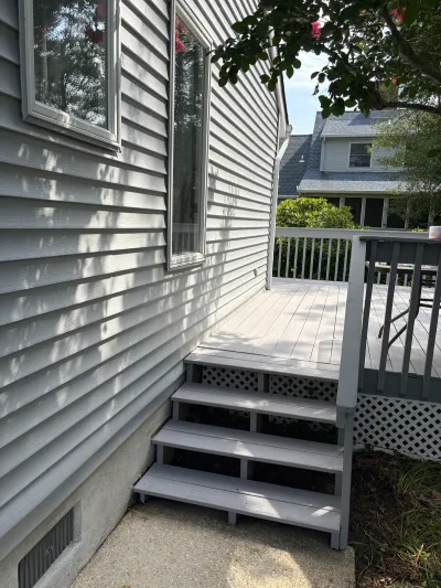 Side view of a house with grey vinyl siding, a wood deck, and stairs leading up to it.