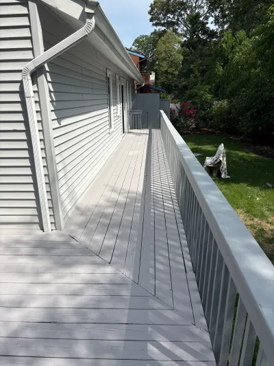 A long side deck with grey painted railing runs alongside a house, overlooking a shaded backyard.