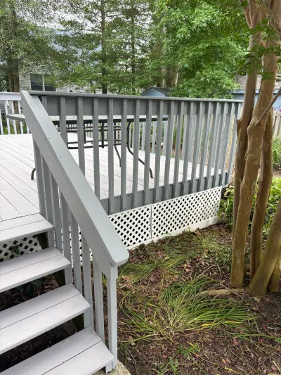 A light grey painted wood deck with stairs, railings, and lattice skirting surrounded by trees.