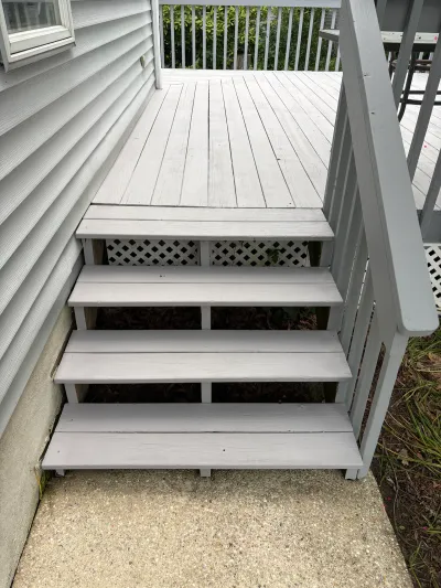 Grey painted wooden deck stairs leading down to a concrete walkway beside a house.