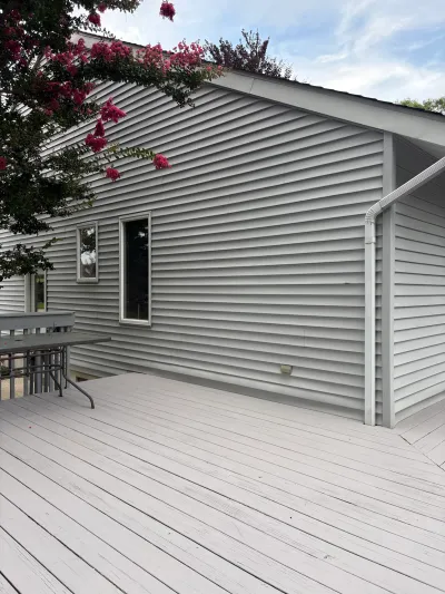 Side view of a house with grey vinyl siding and a wood deck, framed by blooming crepe myrtle branches.