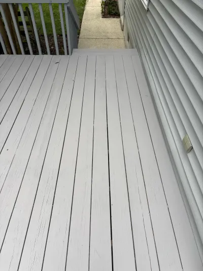 Weathered deck boards with peeling grey paint alongside a house, leading to a concrete walkway.
