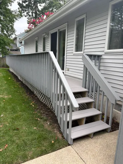 A grey painted deck with railings and stairs runs along the side of a house with vinyl siding.