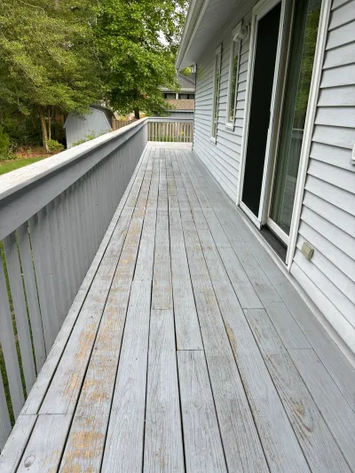 A weathered wood deck with peeling gray paint runs alongside a house with vinyl siding.