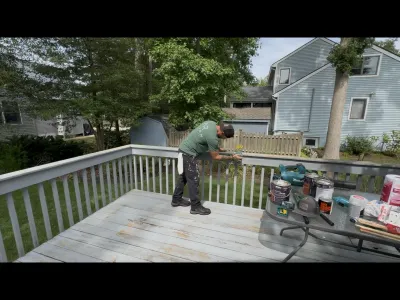 Worker staining a deck railing on a weathered wood deck with paint supplies on a nearby table.