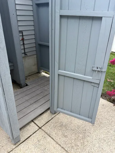 Open door of a gray board and batten outdoor shower enclosure with exposed plumbing inside.