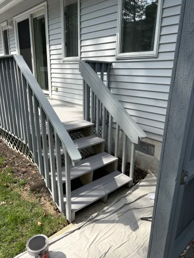 Exterior wooden stairs and railings being repainted gray beside a vinyl-sided house.
