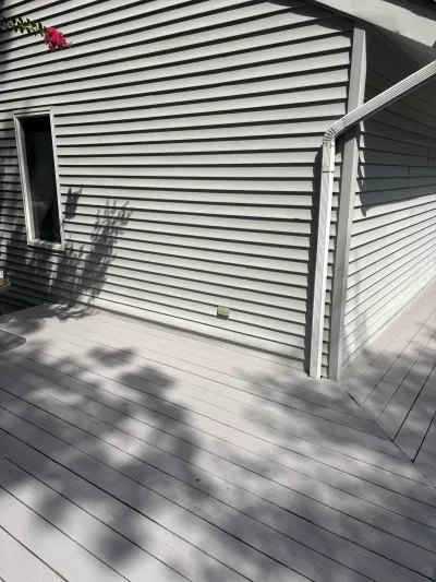 Corner of a house with light grey vinyl siding and a composite deck, dappled in tree shadows.