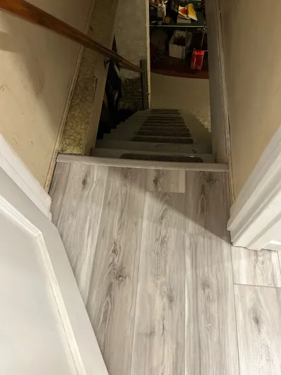 View down a narrow basement stairwell with new wood-look flooring at top transitioning to older carpeted stairs.