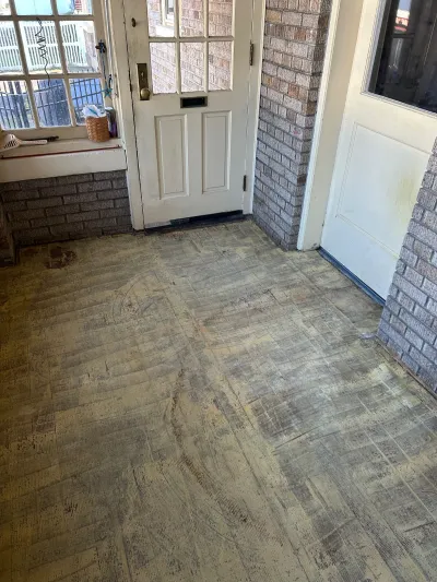 Worn, stained sheet vinyl flooring in an entryway flanked by brick walls and a paneled front door.