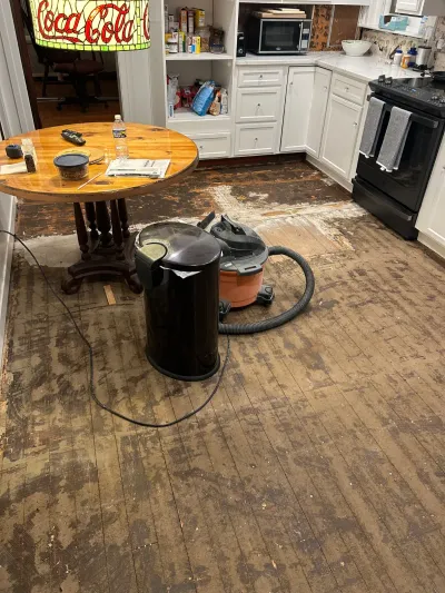 Kitchen with flooring stripped to the subfloor and a shop vacuum mid-remediation, cabinets intact overhead.