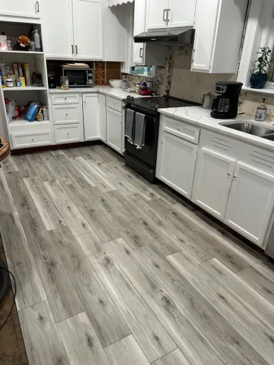 Kitchen mid-renovation with new grey plank flooring and white cabinets, backsplash wall stripped to bare drywall.