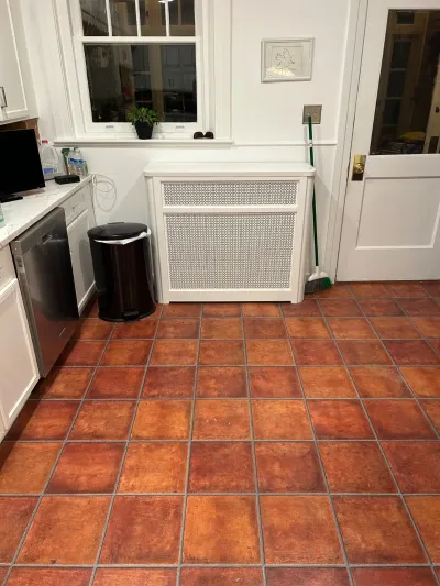 Kitchen corner with terracotta tile floor, white cabinets, and a radiator cover under a window.