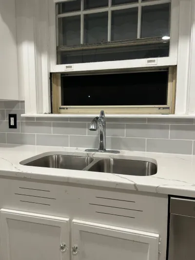 Close-up of a kitchen sink with stainless steel basin, quartz counters, and subway tile backsplash beneath a window.