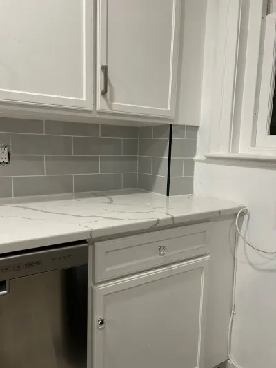 Close-up of a white kitchen corner with quartz counters, grey subway tile backsplash, and a dishwasher.