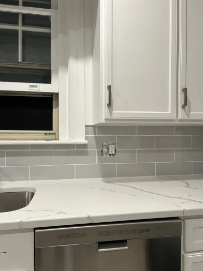 Close-up of a white kitchen with grey subway tile backsplash and quartz countertop by a window.