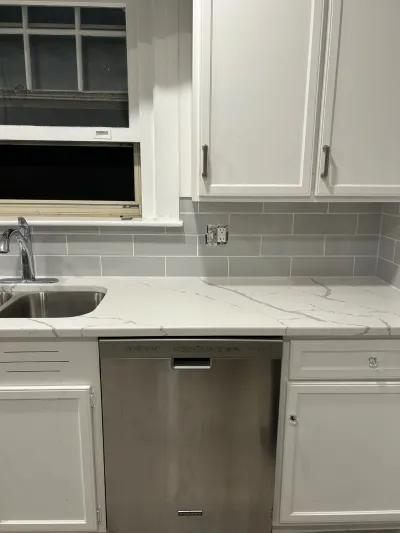 A kitchen counter mid-renovation with a new quartz countertop, subway tile backsplash, and stainless dishwasher.