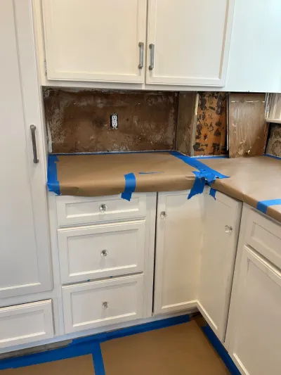Kitchen backsplash removed down to drywall, with countertops and floor taped off for protection during renovation.