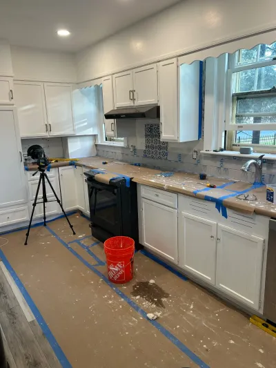Kitchen mid-renovation with white cabinets, protective paper on counters and floor, and partially tiled backsplash.