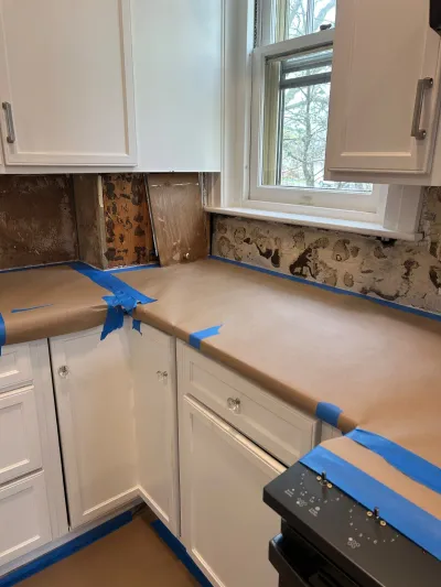 Kitchen mid-renovation with backsplash removed and countertops covered in protective paper and tape.