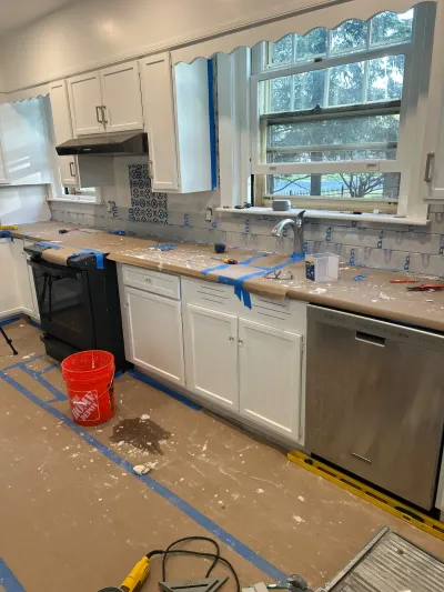 A kitchen mid-remodel with white cabinets, partially tiled backsplash, and protected countertops and flooring.