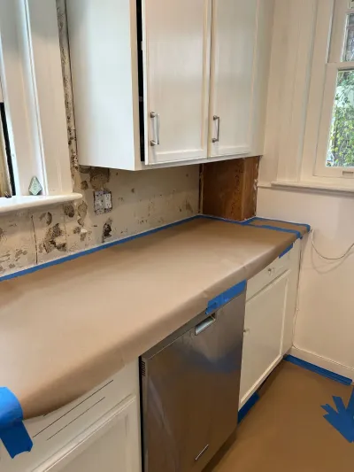 Kitchen mid-renovation with taped-off counters, stripped backsplash, and white cabinetry.
