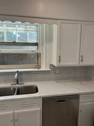 Kitchen sink area with quartz countertop, subway tile backsplash, and white shaker cabinets beside a window.