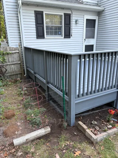 Grey painted deck attached to a white vinyl-sided house with a garden bed and tomato cages below.