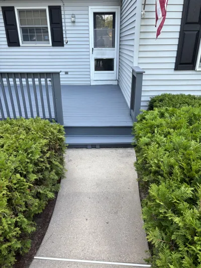 Front porch with grey painted deck, white storm door, and concrete walkway lined by trimmed shrubs.