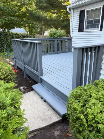 A newly painted grey wood deck with railings and steps leading to a concrete walkway beside a house.