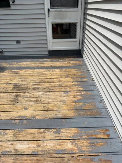 A worn wood deck with peeling grey paint next to a white vinyl-sided house and exterior door.