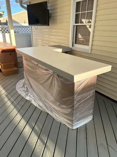 An outdoor bar counter wrapped in plastic sits on a deck beneath a mounted TV during installation.