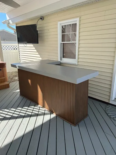 Outdoor bar counter with wood paneled base and grey countertop on a covered deck, with a mounted TV above.