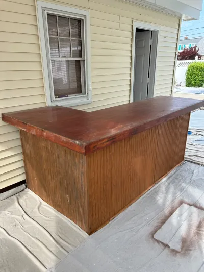 An outdoor built-in bar with a stained wood top and beadboard sides under construction beside a house.