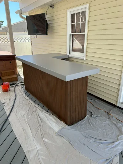 An L-shaped outdoor bar with a gray countertop and wood paneling under construction on a covered deck.