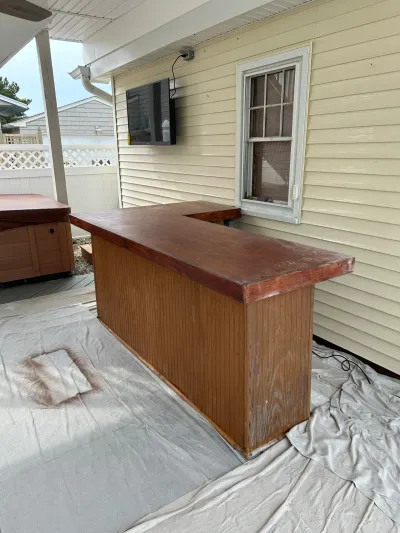 Outdoor L-shaped wood bar with freshly stained top, drop cloths on the patio floor below.